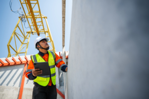 Male civil engineer in safety workwear using a digital tablet for site inspection while looking up at a heavy yellow gantry crane at an industrial precast concrete construction project.